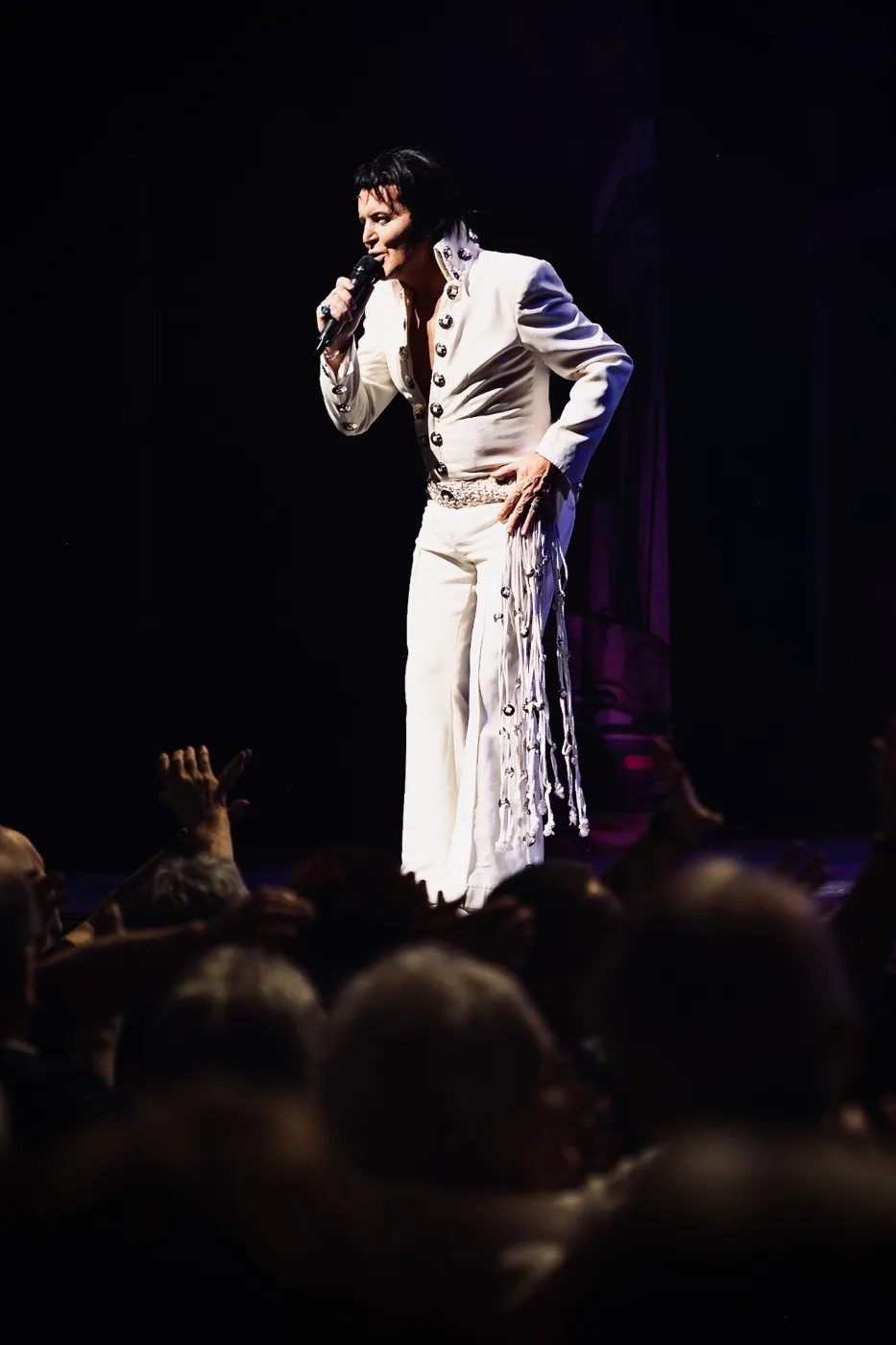 A performer in a white fringed stage suit singing into a microphone at the front of the stage, with audience members raising their hands below