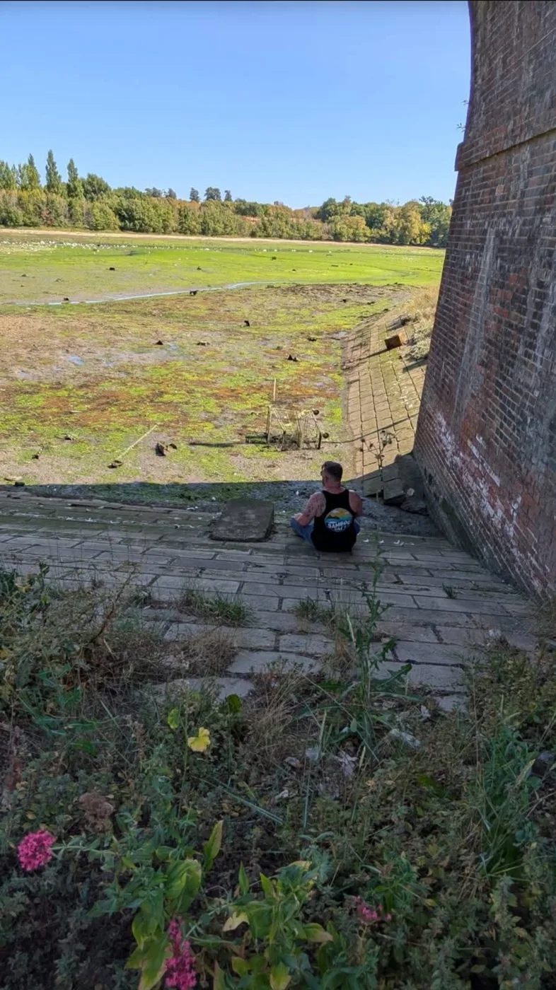 Lee Knight sitting on the stone slope beside a brick pier of the railway bridge at Fareham Creek, looking out over the creek bed